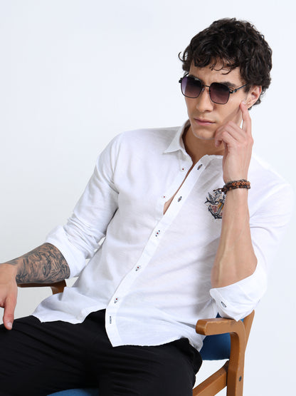 Man wearing a white shirt with a patterned design, sitting on a chair against a plain background.