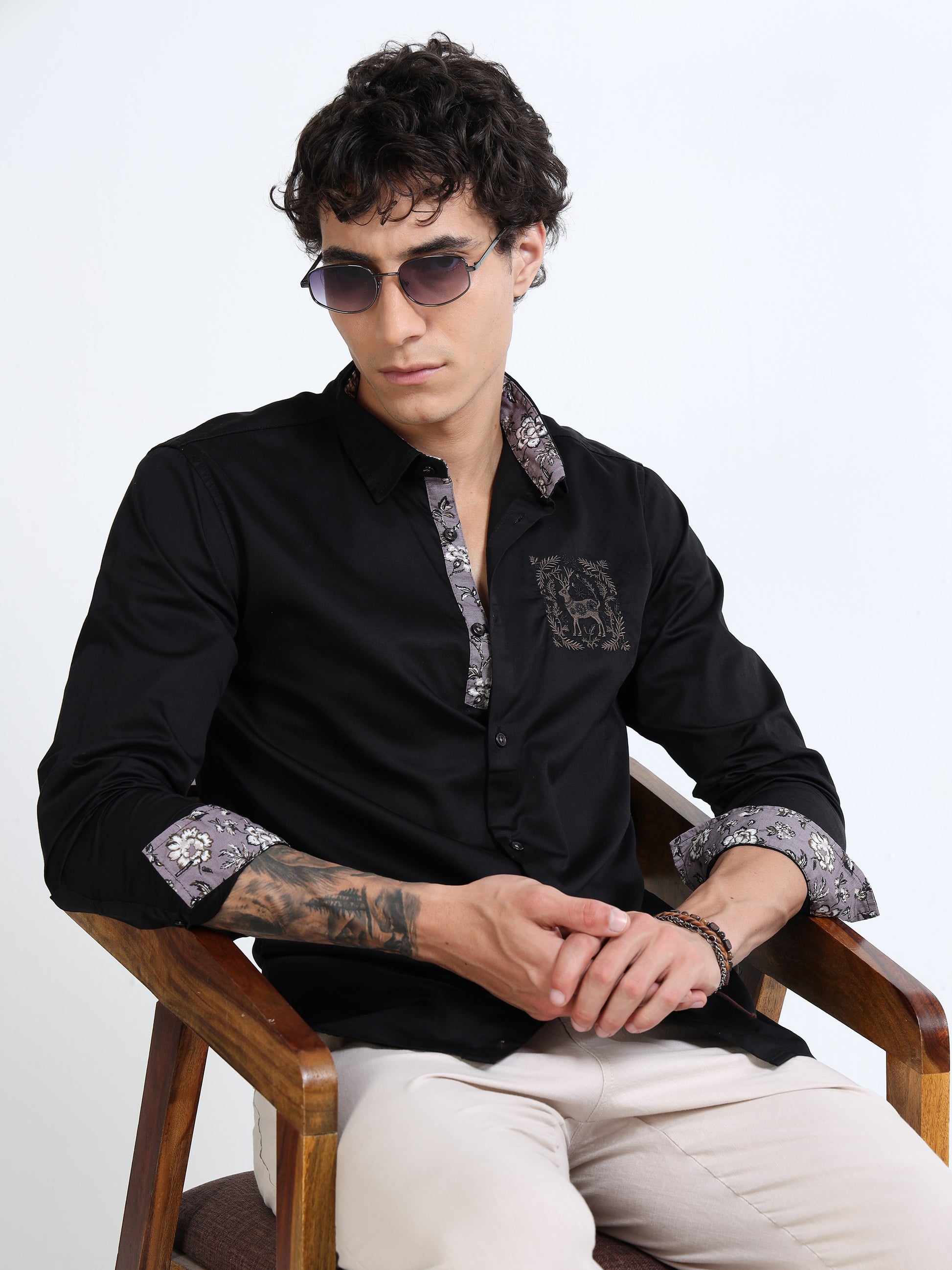 Man wearing a black shirt with patterned cuffs and sunglasses, sitting on a wooden chair against a white background.