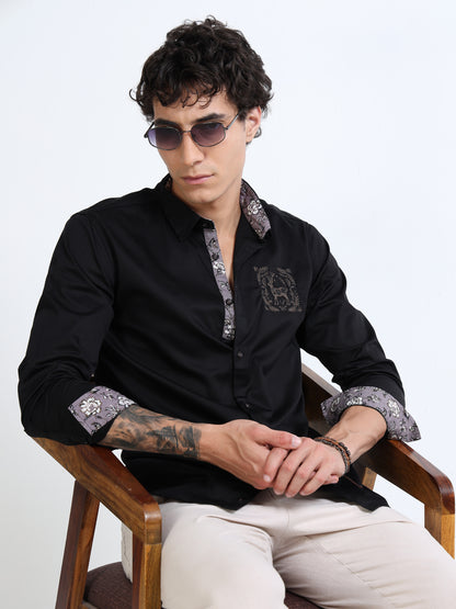 Man wearing a black shirt with patterned cuffs and sunglasses, sitting on a wooden chair against a white background.