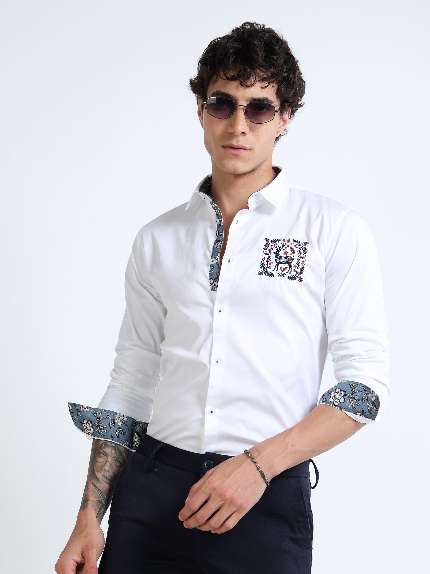 Man wearing a white dress shirt with floral cuffs and collar, sunglasses, and dark pants on a white background