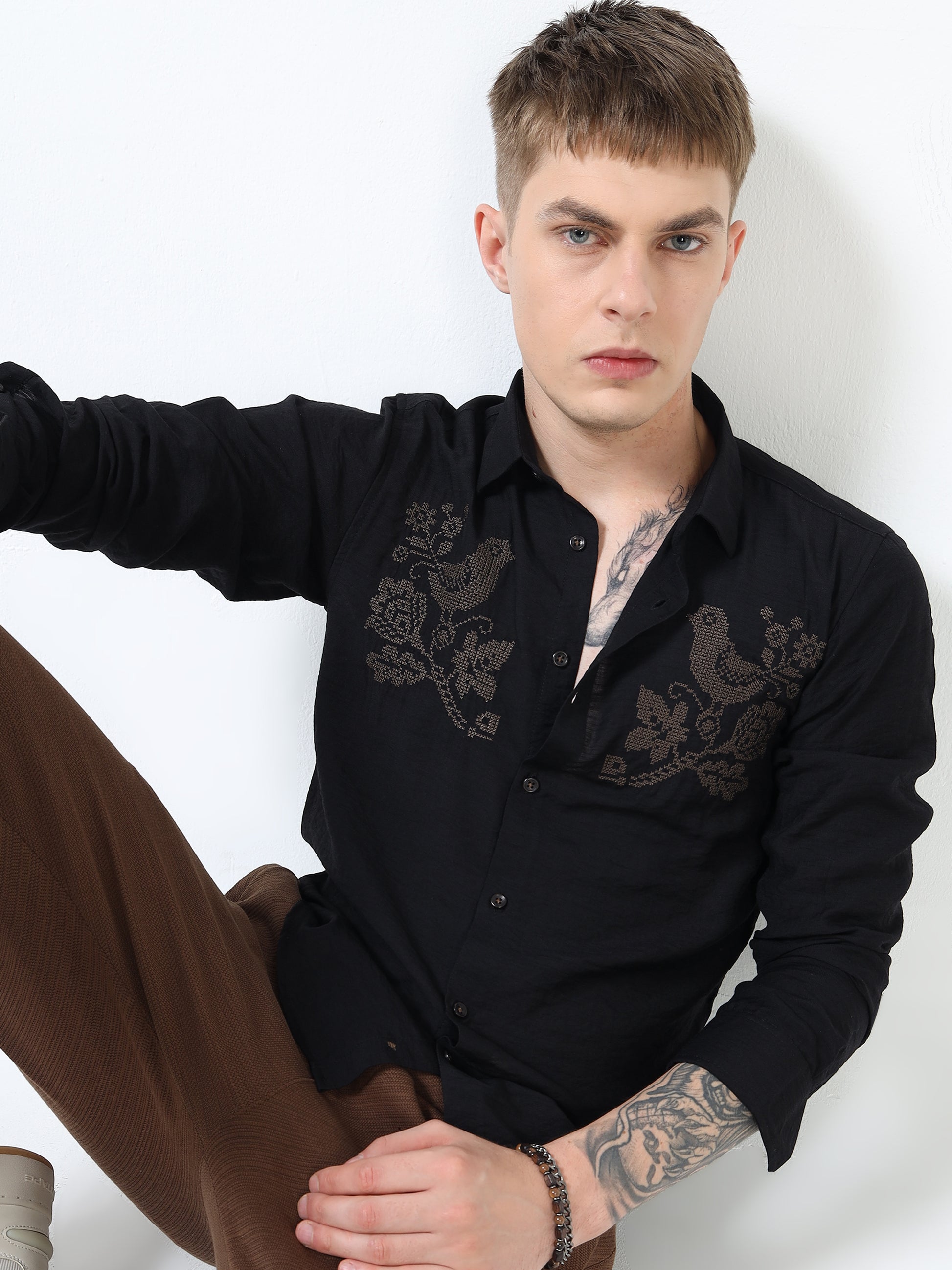 Man wearing a black embroidered shirt sitting against a white background