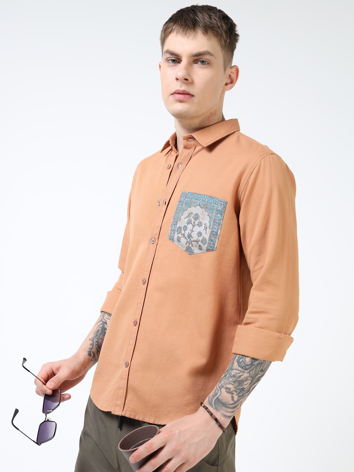Man wearing a brown shirt with a patterned pocket, holding sunglasses, on a white background