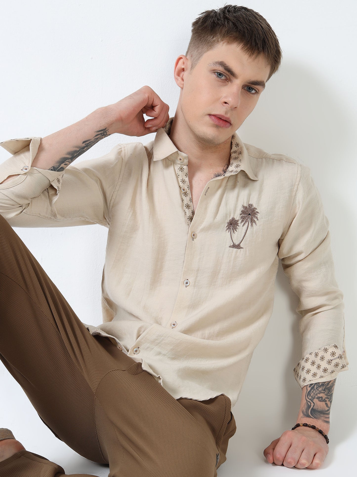 Man wearing a beige shirt with a palm tree design and brown pants against a white background