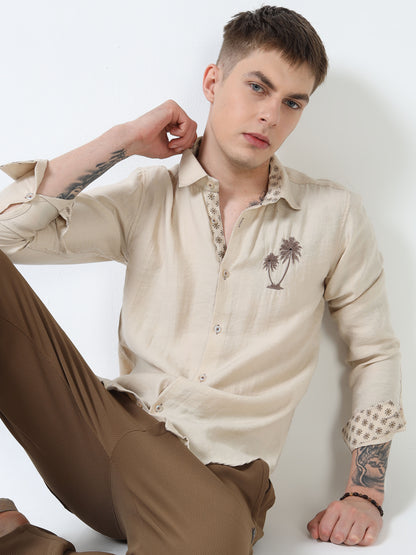 Man wearing a beige shirt with a palm tree design and brown pants against a white background